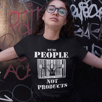 Woman standing against a graffiti wall, wearing a 'we're people not products' t shirt. An anti capitalism t shirt highlighting the dehumanisation of people in a money obsessed society. A fantastic choice for any anti capitalism protest or to just create social awareness. Made from 100% organic ring-spun cotton, this anti consumerism t shit is unisex, high-quality, super comfy, and best of all, eco-friendly.