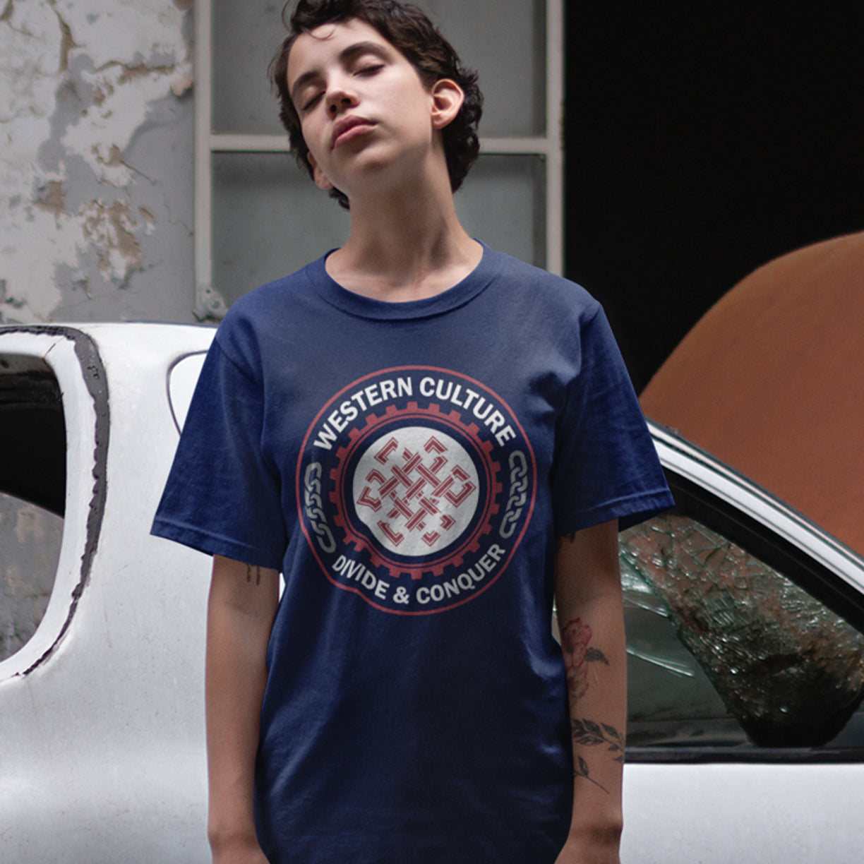 Person wearing a French navy blue t-shirt with a graphic design in front of a car. This counter culture t shirt questions the politics of western society. Let's drop the façade that western culture is more civilised and advanced. This design highlights a method of people management that has worked for the Western elite for centuries. Made from 100% organic ring-spun cotton, this broken society t shirt is unisex, high-quality, super comfy, and best of all, eco-friendly.