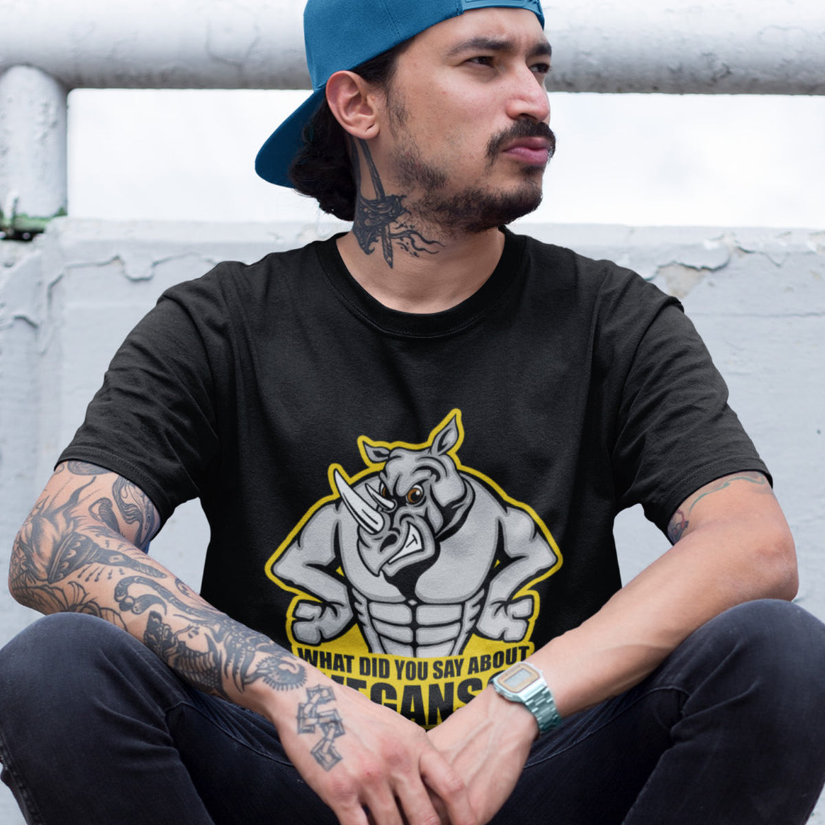 A man in a blue cap, sitting on a step, wearing a black short sleeved crew neck t-shirt with a cartoon Rhino design and the caption "WHAT DID YOU SAY ABOUT VEGANS?".