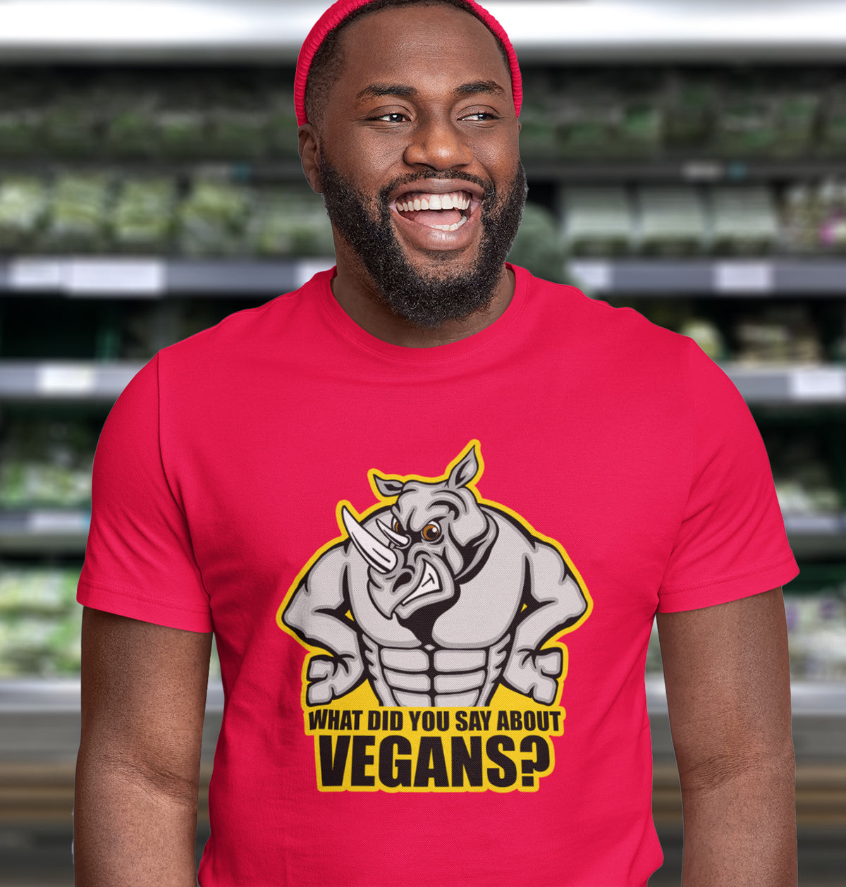 A laughing man standing in a supermarket, wearing a beany hat and a red short sleeved crew neck t-shirt with a cartoon Rhino design and the caption "WHAT DID YOU SAY ABOUT VEGANS?".