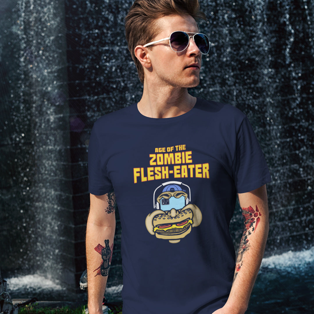 Man standing in front of a waterfall wearing a French navy blue t-shirt with a graphic on the front. The graphic features the text 'AGE OF THE ZOMBIE FLESH-EATER' in yellow, above a cartoon zombie head wearing headphones and biting a burger.