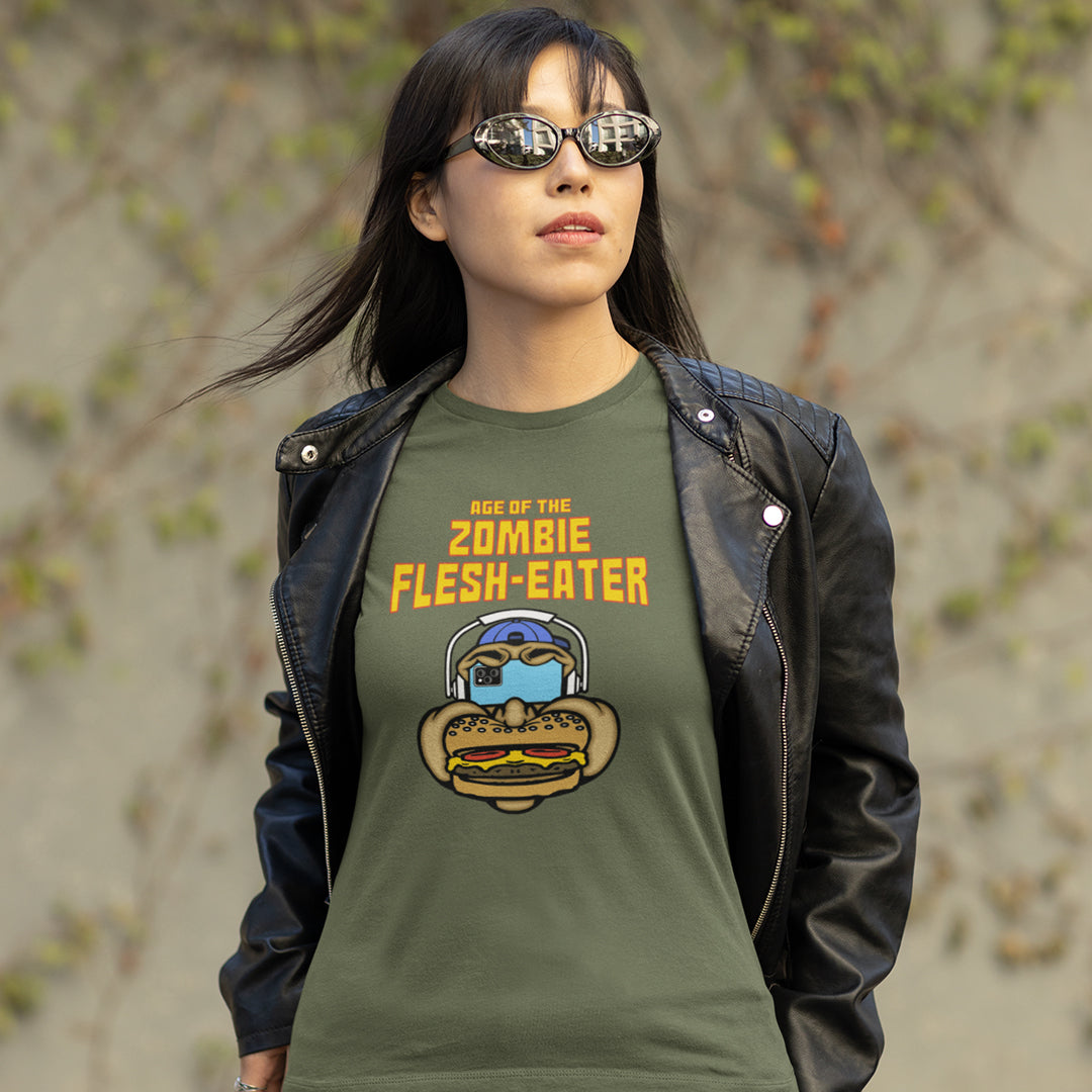 A woman wearing a khaki t shirt with a graphic on the front. The graphic features the text 'AGE OF THE ZOMBIE FLESH-EATER' in yellow, above a cartoon zombie head wearing headphones and biting a burger.