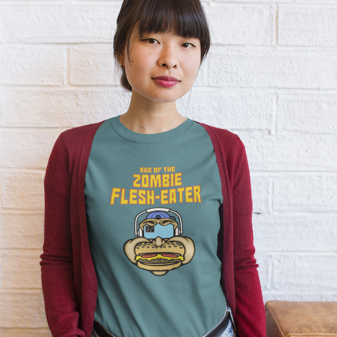 A woman standing in front of a brick wall wearing a stargazer blue t-shirt with a graphic on the front. The graphic features the text 'AGE OF THE ZOMBIE FLESH-EATER' in yellow, above a cartoon zombie head wearing headphones and biting a burger.