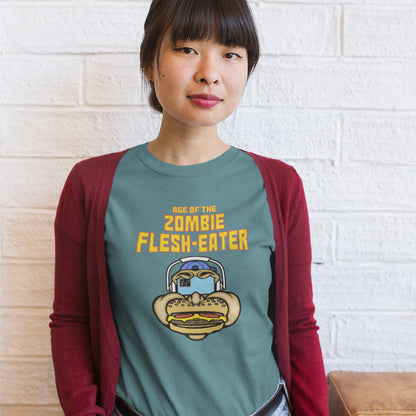A woman standing in front of a brick wall wearing a stargazer blue t-shirt with a graphic on the front. The graphic features the text 'AGE OF THE ZOMBIE FLESH-EATER' in yellow, above a cartoon zombie head wearing headphones and biting a burger.